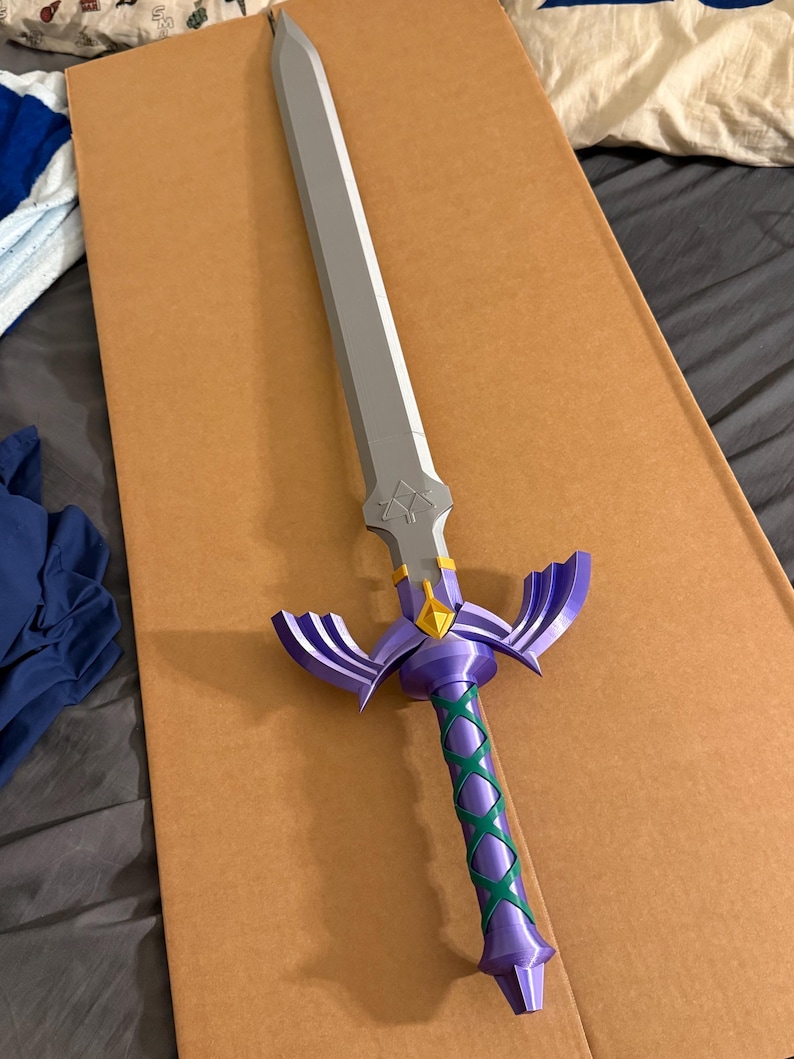 May include: A decorative sword with a silver blade and a purple hilt. The hilt features a green crisscross pattern and purple wings. A gold diamond shape is at the base of the blade. The sword is lying on a brown surface.