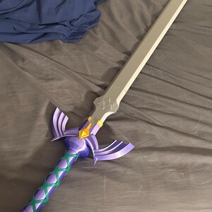 May include: A decorative sword with a silver blade and a purple hilt. The hilt features a green crisscross pattern and a gold accent. The sword is lying on a gray surface.