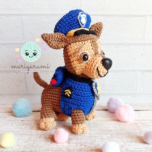 May include: A crocheted Chase the police dog toy from the Paw Patrol series. The toy is brown and tan with a blue police uniform and hat. The toy has a black nose and a smiling expression. The logo "marigurumi" is in the image.