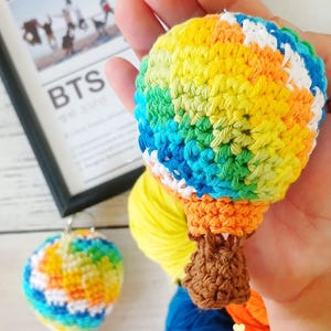 Puede incluir: Dos globos aerostáticos de crochet coloridos, uno en una mano y otro en una mesa, con un fondo de ovillos de lana y una foto enmarcada de una banda de chicos con el texto "BTS".