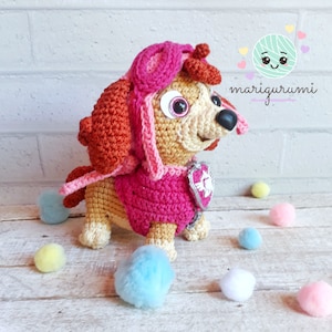 May include: A crocheted plush toy dog wearing a pink and orange costume with a silver badge. The dog has brown fur, pink ears, and a pink nose. The toy is sitting on a wooden surface with white, pink, and blue pom-poms.