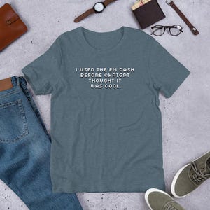 Può includere: Una maglietta color verde acqua con il testo pixelato "I USED THE EM DASH BEFORE CHATGPT THOUGHT IT WAS COOL." La maglietta è esposta su una superficie piana con jeans, scarpe, un orologio e altri accessori.