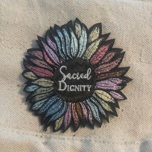 May include: Embroidered patch in the shape of a sunflower with petals in shades of blue, pink, purple, and gold. The center is black with the words "Sacred Dignity" embroidered in white.