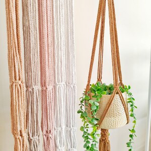 Op de afbeelding: Een collectie macramé plantenhangers in tinten beige, roze en wit, met geknoopte koordontwerpen. Eén bruine hanger bevat een metallic gouden gehamerde pot met een hangende kunstplant met kleine ronde blaadjes.
