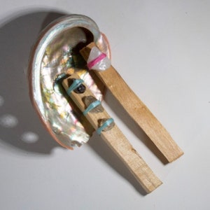 May include: A seashell with iridescent colors holds two wooden sticks with crystals and rubber bands. The crystals are clear, pink, and black.