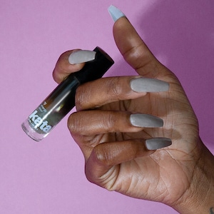 Puede incluir: Una botella de vidrio transparente con una tapa negra y una etiqueta blanca que dice "kata". La botella está llena de un líquido oscuro. La botella está siendo sostenida por una mano con esmalte de uñas gris.