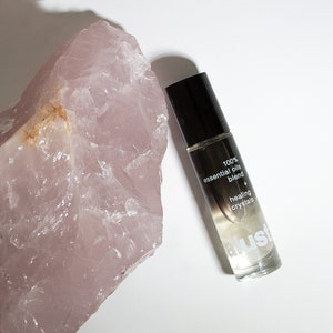 May include: A clear glass bottle with a black cap and white label. The label reads "100% essential oils blend + healing crystals" and "alustr". The bottle is sitting on a pink rose quartz crystal.