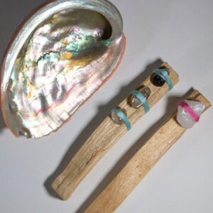 Może przedstawiać: Dwa drewniane patyczki z kryształami i sznurkiem owiniętymi wokół. Patyczki leżą na białym tle. Na zdjęciu znajduje się również duża, tęczowa muszla abalonu.