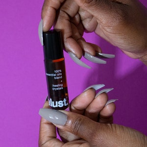 May include: A small brown glass bottle with a black roller ball top. The bottle is labeled with white text that reads "100% essential oils blend + healing crystals lust."