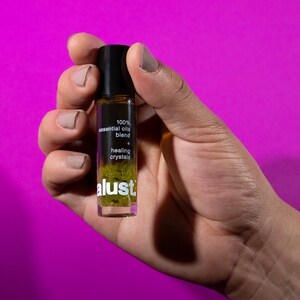 Puede incluir: Un frasco de aceite esencial en rollo de vidrio transparente con tapa negra, sobre un fondo magenta. El frasco contiene una mezcla de aceites esenciales y cristales curativos, con el texto "100% essential oils blend + healing crystals" y la marca "alust".
