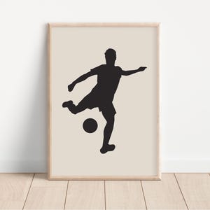 May include: A framed print featuring a black silhouette of a soccer player kicking a ball. The player is in mid-action, with one leg extended towards the ball. The background is a neutral beige color, and the frame is a light wood tone.