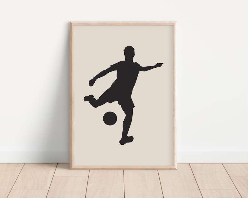 May include: A framed print featuring a black silhouette of a soccer player kicking a ball. The player is in mid-action, with one leg extended towards the ball. The background is a neutral beige color, and the frame is a light wood tone.