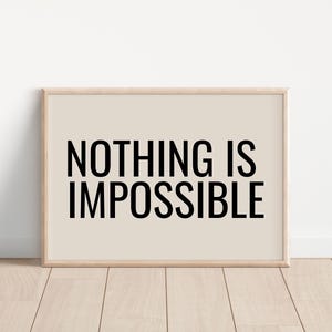 May include: A framed print with the words "NOTHING IS IMPOSSIBLE" in bold black letters. The background is a light beige color, and the frame is a light wood color. The print is displayed on a wooden floor.