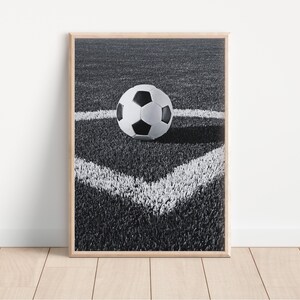 May include: Framed black and white print of a soccer ball on a green field. The ball is positioned in the corner of the field, marked by white lines. The image is set in a wooden frame and placed against a white wall.