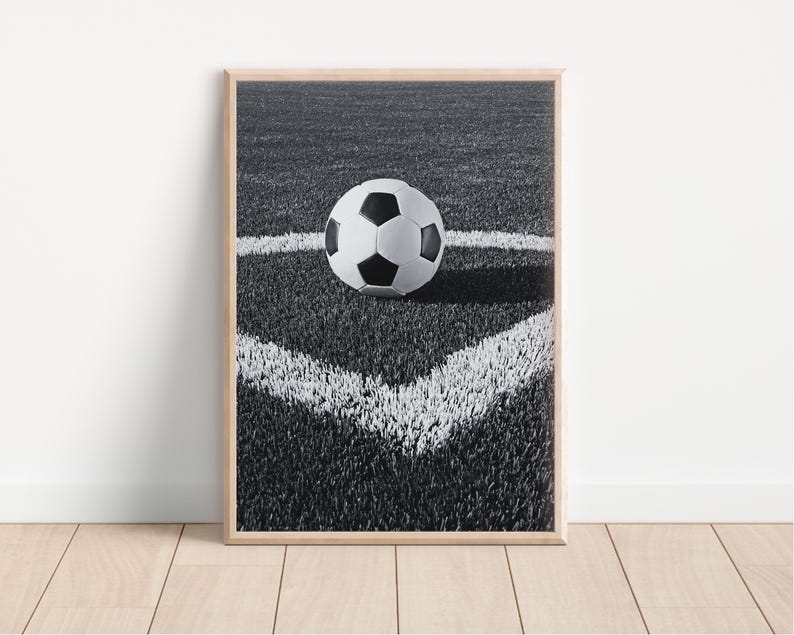 May include: Framed black and white print of a soccer ball on a green field. The ball is positioned in the corner of the field, marked by white lines. The image is set in a wooden frame and placed against a white wall.