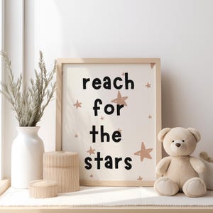 May include: A framed print with the text "reach for the stars" in black lettering on a cream background with brown star illustrations. A teddy bear sits on a table in front of the print.
