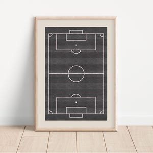 May include: Framed print of a soccer field, featuring a black and gray background with white lines marking the field's boundaries. The print is encased in a light-colored wooden frame, suitable for sports enthusiasts and home decor.