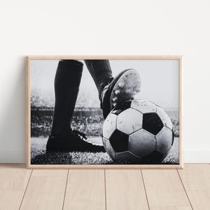 May include: Black and white framed print of a soccer ball with a player's foot on top. The soccer ball is on a grassy field, and the player is wearing cleats and a uniform. The background shows a stadium.