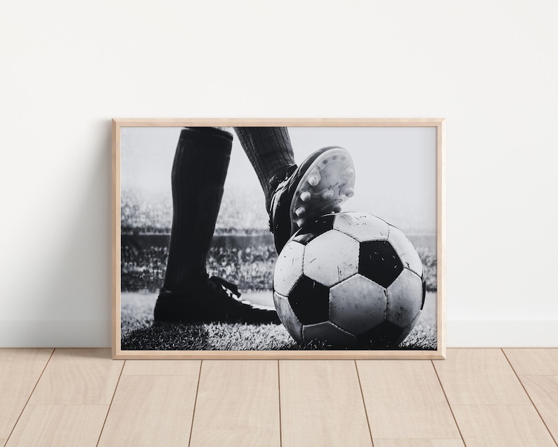 May include: Black and white framed print of a soccer ball with a player's foot on top. The soccer ball is on a grassy field, and the player is wearing cleats and a uniform. The background shows a stadium.