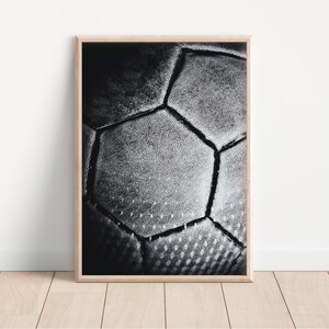 May include: Black and white framed print featuring a close-up of a soccer ball. The image highlights the textured surface and geometric pattern of the ball, with visible stitching. The print is displayed in a light-colored wooden frame.
