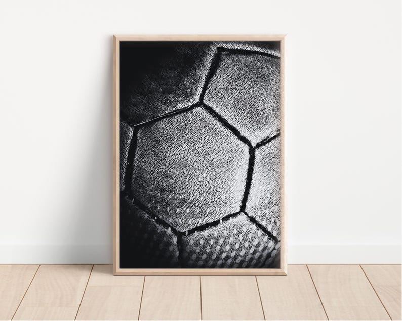 May include: Black and white framed print featuring a close-up of a soccer ball. The image highlights the textured surface and geometric pattern of the ball, with visible stitching. The print is displayed in a light-colored wooden frame.