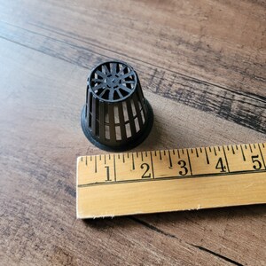 May include: A black plastic net pot with a circular top and vertical bars. The pot is approximately 2 inches in diameter. A wooden ruler is next to the pot.