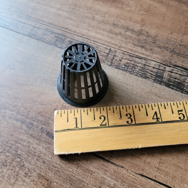 May include: A black plastic net pot with a circular top and vertical bars. The pot is approximately 2 inches in diameter. A wooden ruler is next to the pot.