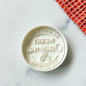 May include: A white, circular cookie cutter with the words "Merry Christmas" and "We Wish You A Joyful Christmas" in a decorative font. The design includes holly leaves. A red and white patterned cloth is in the background.