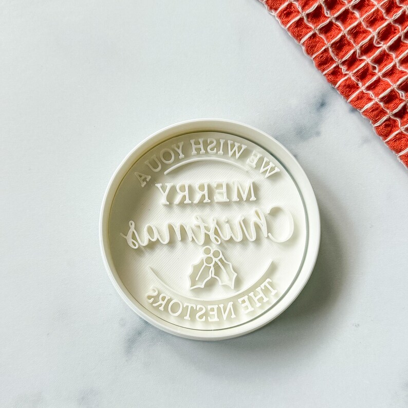 May include: A white, circular cookie cutter with the words "Merry Christmas" and "We Wish You A Joyful Christmas" in a decorative font. The design includes holly leaves. A red and white patterned cloth is in the background.