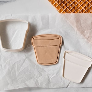 May include: Two cookie cutters in the shape of a flower pot. One cutter is white and the other is brown. The cutters are on a white surface.