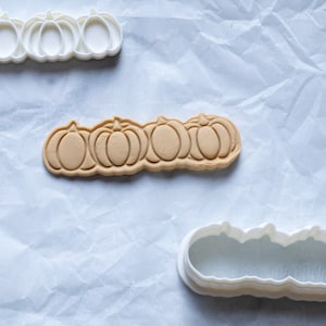 May include: An overhead shot displays a pumpkin-shaped cookie cutter and a baked cookie. The cookie cutter is white, and the baked cookie is light brown. The cookie cutter and cookie are arranged on a white, textured surface.