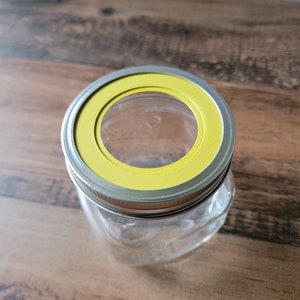 May include: A clear glass jar with a silver metal lid and a bright yellow ring insert. The jar is empty and sits on a wooden surface. The jar is square with rounded edges.