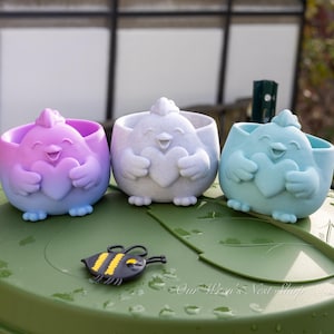 May include: Three colorful, smiling chick-shaped planters, each holding a heart. The planters are in shades of purple, white, and blue. A small bee-shaped charm rests on a green surface in front of the planters. The planters are likely for small plants or succulents.