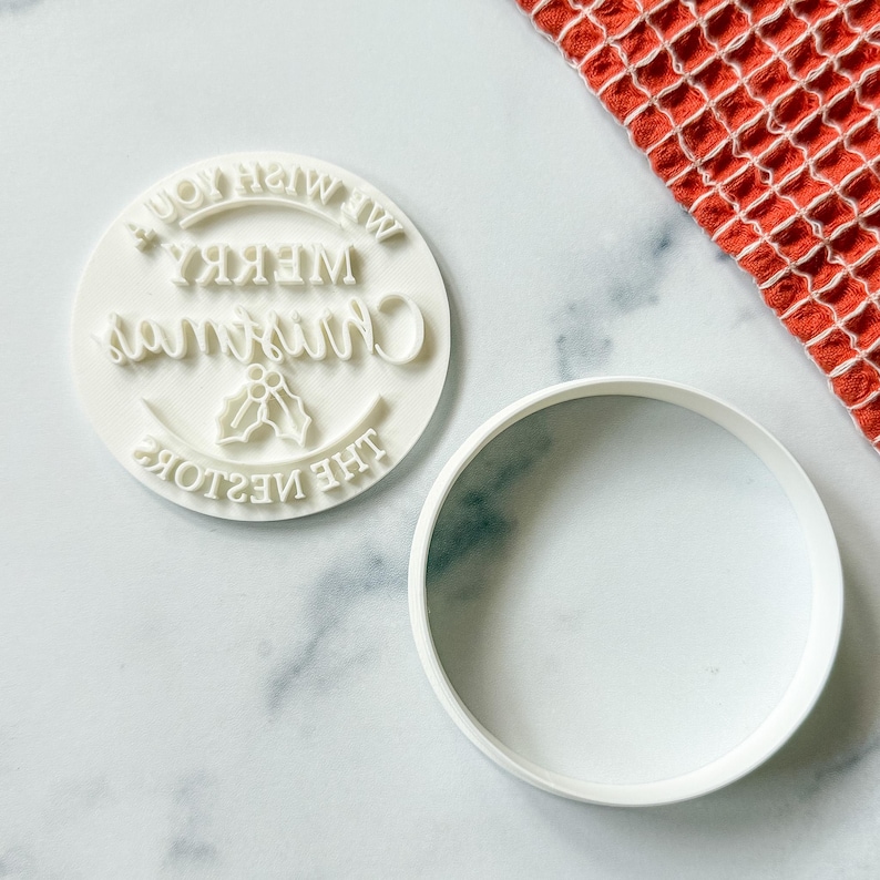 May include: A white, round cookie stamp with the words "Merry Christmas" and "We Wish You" in a circular design. The stamp includes a holly sprig and a separate white, round cookie cutter. A red and white patterned cloth is in the background.