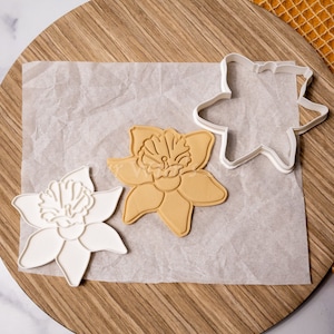 May include: Two white plastic cookie cutters and a yellow cookie in the shape of a daffodil flower on a brown wooden surface.