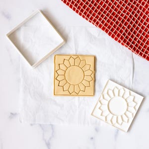 May include: Two white plastic cookie cutters, one shaped like a square and the other shaped like a sunflower, are on a white surface. A piece of cookie dough is pressed into the sunflower cookie cutter.