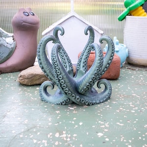 May include: A detailed, teal-colored octopus sculpture with curled tentacles. The sculpture has a textured surface and is positioned on a green surface. Other garden decor items are visible in the background.