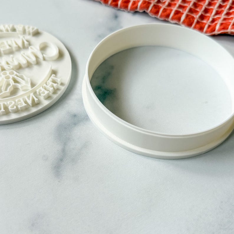 May include: A white, round cookie cutter and a matching circular stamp. The stamp has the words "Merry Christmas" and holly leaves. The cutter is a ring shape. The items are on a white marble surface.
