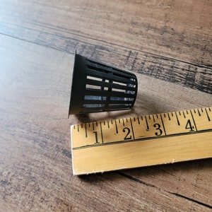 May include: A black plastic net pot with slotted sides, used for hydroponics or container gardening. The pot is approximately 2 inches in diameter and sits next to a wooden ruler.