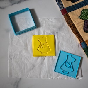 May include: A set of square cookie cutters and a matching yellow square stamp featuring a silhouette of a person wearing a bonnet and a dress. The set includes one empty blue square frame, one blue square stamp, and one yellow square stamp.