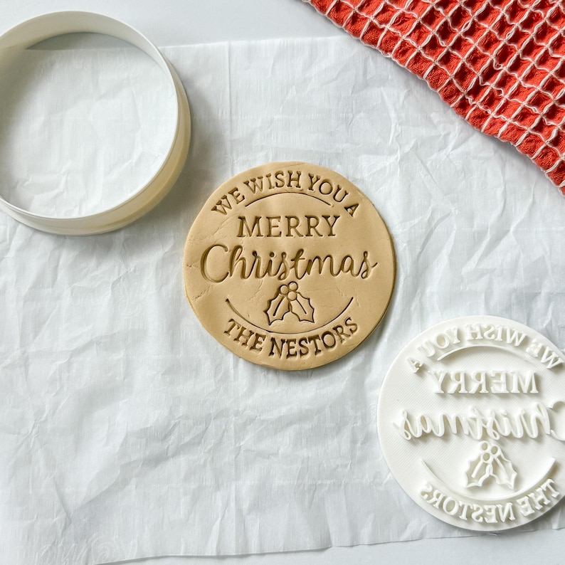 May include: A round cookie with the words "WE WISH YOU A MERRY Christmas" and "THE NESTORS" imprinted on it. A white cookie cutter and stamp are also visible. A red and white checkered cloth is in the background.