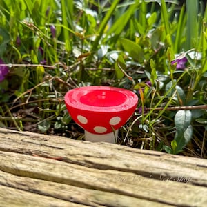 May include: A red mushroom-shaped decorative item with white polka dots. The mushroom has a rounded top and a white stem. The item is set outdoors among green foliage and a wooden surface.
