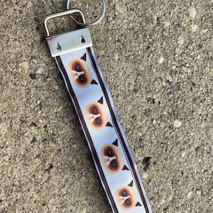 May include: A white and light blue patterned keychain wristlet featuring a repeating design of a cat's face with blue eyes and brown accents. The wristlet is attached to a silver-colored metal clasp and keyring.