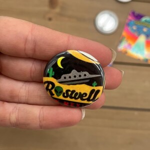Peut inclure: Un bouton rond avec un fond noir et un design jaune, orange et vert. Le bouton repr&eacute;sente un OVNI de dessin anim&eacute;, une lune croissante et le texte "Roswell, Nouveau-Mexique".