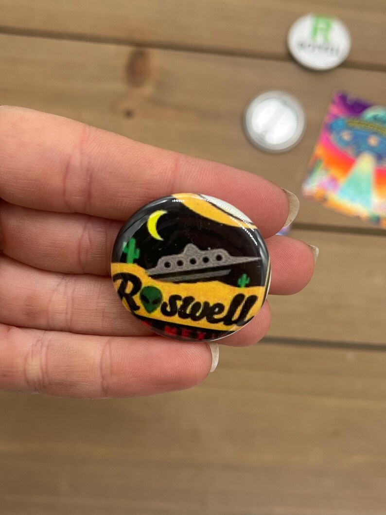 Peut inclure: Un bouton rond avec un fond noir et un design jaune, orange et vert. Le bouton repr&eacute;sente un OVNI de dessin anim&eacute;, une lune croissante et le texte "Roswell, Nouveau-Mexique".