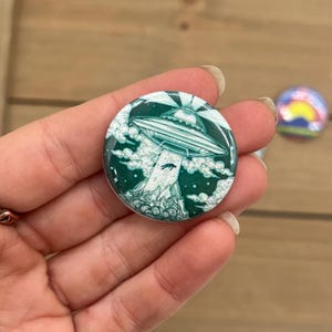 Peut inclure: Un bouton vert et blanc avec une image de dessin anim&eacute; d'un OVNI volant au-dessus d'un paysage vert et blanc.