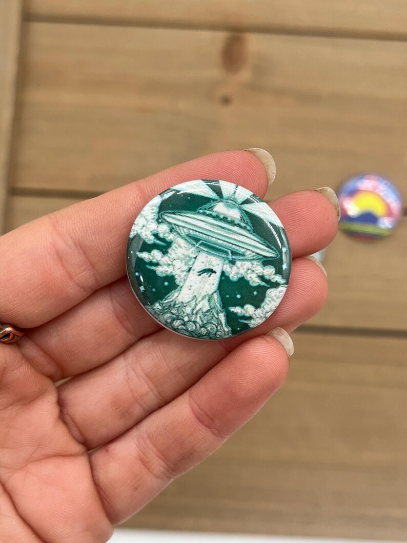 Peut inclure: Un bouton vert et blanc avec une image de dessin anim&eacute; d'un OVNI volant au-dessus d'un paysage vert et blanc.