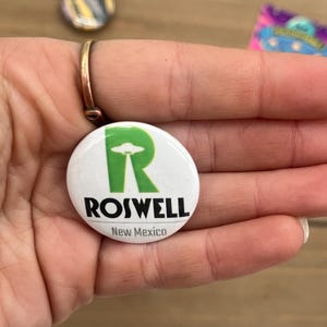 Peut inclure: Bouton blanc avec un "R" vert et les mots "Roswell, Nouveau-Mexique". Le "R" contient un OVNI vert.