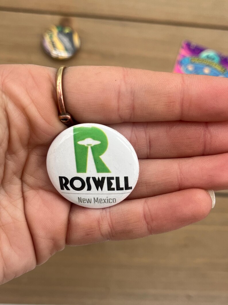 Peut inclure: Bouton blanc avec un "R" vert et les mots "Roswell, Nouveau-Mexique". Le "R" contient un OVNI vert.