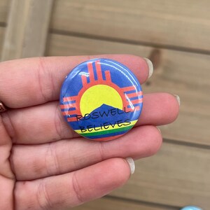 Peut inclure: Un bouton rond avec un fond bleu et un soleil jaune au centre. Le soleil est entour&eacute; d'un symbole rouge et orange. Le texte "Roswell Believes" est &eacute;crit en noir sous le symbole.
